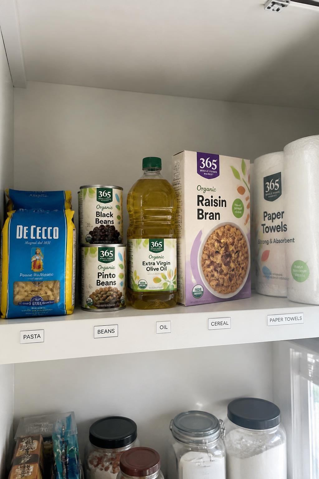 Image prompt: Upper pantry shelf holding a controlled amount of grocery backstock including two extra pasta packets, spare canned beans, cooking oil, cereal, and paper towels, each category kept behind one small label, photographed with modern iPhone photo quality, natural window light, realistic household quantities, simple clean pantry, no excessive bulk products, no people, no watermarks on images.