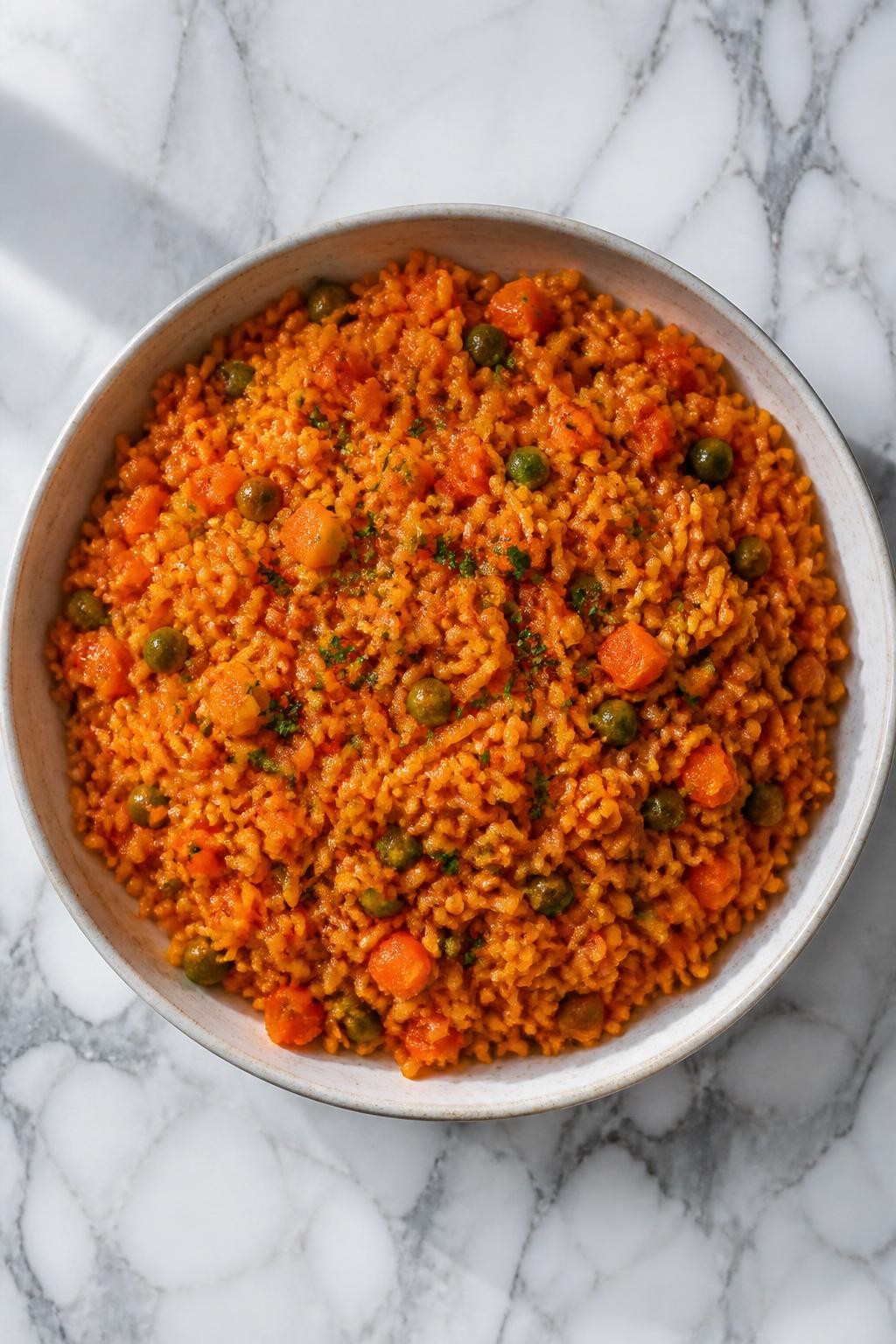 Image Prompt: Top-down realistic editorial food photography of a bowl of vegan jollof rice on a clean white and gray marble countertop, fluffy orange-red rice cooked in rich tomato and pepper sauce with carrots, green peas, and herbs, bright natural daylight, warm homemade African dinner mood, clean composition, high detail, realistic food texture, no people, no hands, no text, no watermarks, no props with writing.