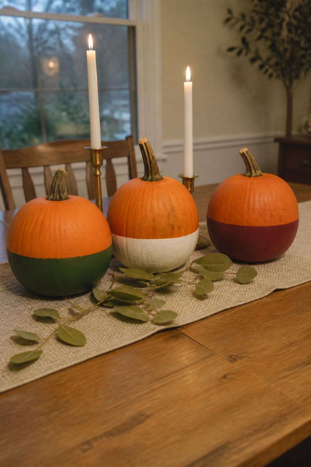 Horizontal 16:9 realistic iPhone photograph of three real orange pumpkins with only their lower halves painted in matte forest green, warm white, and deep burgundy, slightly varied dividing lines, arranged on a simple wooden dining table with a beige runner, taper candles, and loose eucalyptus leaves, soft evening window light, authentic home styling, natural pumpkin blemishes, no professional product photography, no text.