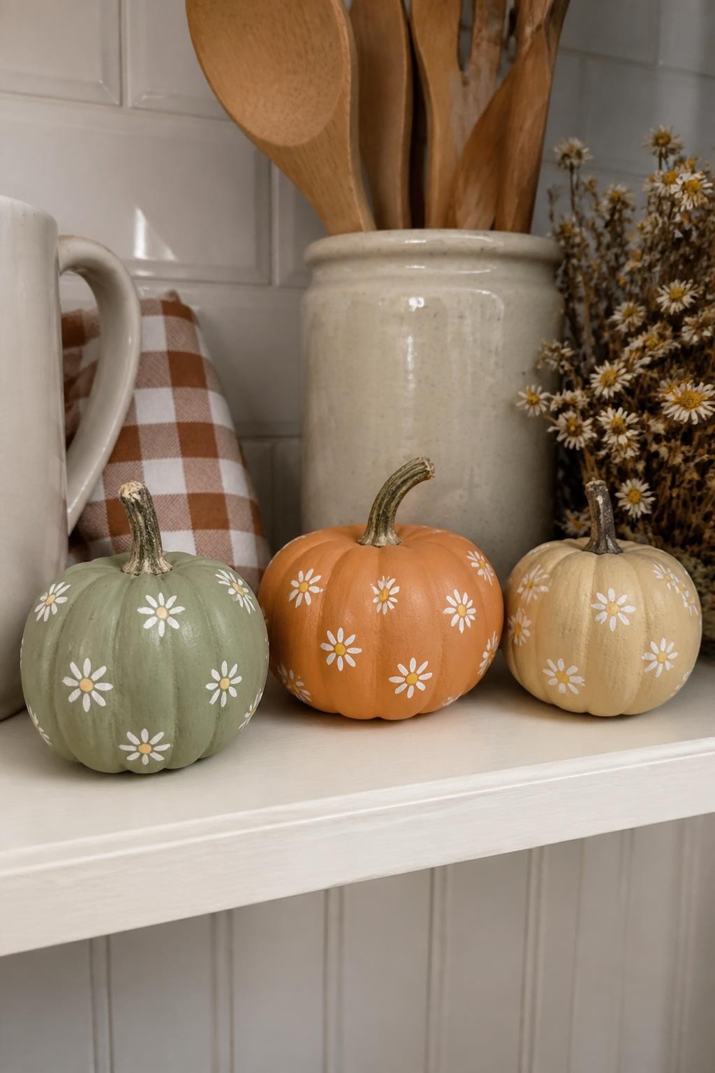 Horizontal 16:9 realistic iPhone photo of three real mini pumpkins painted muted sage, pale orange, and warm beige, each decorated with scattered tiny white daisies with mustard yellow centers, slightly uneven hand-painted petals, displayed on a small kitchen shelf beside a ceramic mug, folded checked napkin, wooden spoon jar, and dried chamomile flowers, soft morning daylight, natural home setting, authentic brush texture, no glossy plastic appearance, no text.