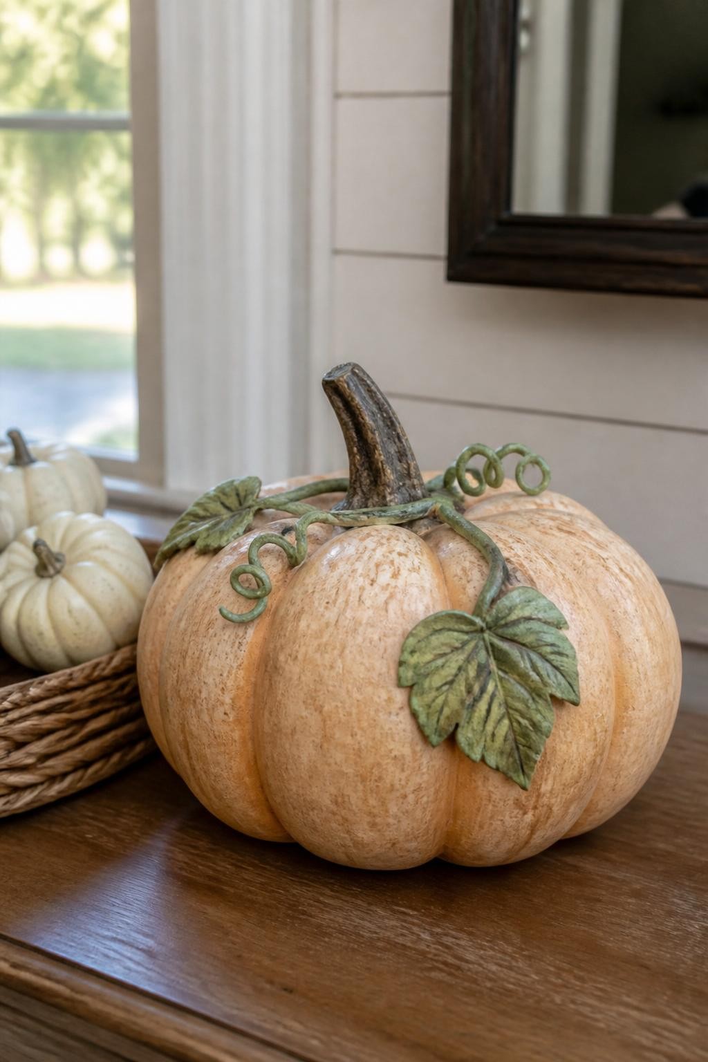 Horizontal 16:9 realistic iPhone photo of a pale orange faux pumpkin with a carefully repainted rough brown stem, muted green vine curling naturally across the top, two realistic painted leaves, gentle rust shading in the grooves, displayed on an entryway table beside a woven tray and small cream pumpkins, soft morning window light, authentic handcrafted detail, no plastic shine, no text.
