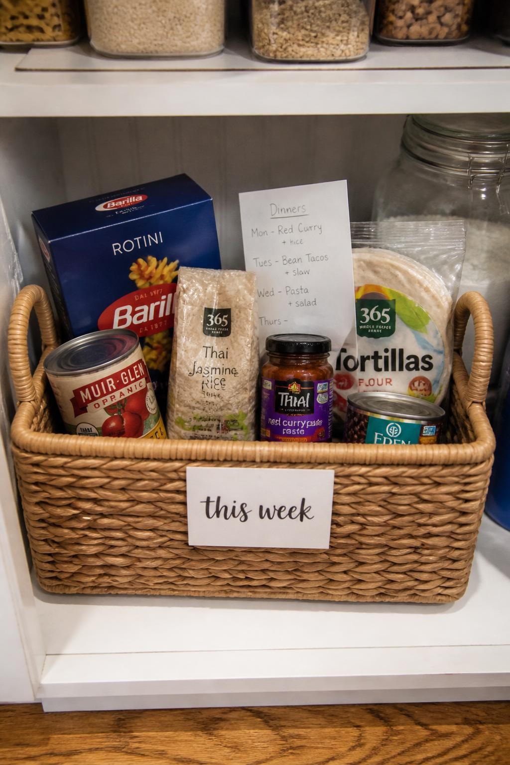 Image prompt: Pantry shelf with one large handled basket labeled “this week,” containing pasta, canned tomatoes, rice, tortillas, beans, curry paste, and recipe notes for planned dinners, photographed with natural iPhone photo quality, bright evening kitchen light, believable mixed grocery packaging, slightly informal home organization, no people, no distorted lettering, no watermarks on images.