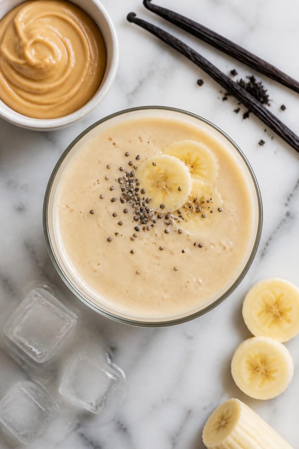 Realistic top-down editorial food photography of a creamy peanut butter vanilla smoothie in a clear glass on a light marble countertop. The smoothie is pale cream-tan, thick, and smooth, styled with peanut butter in a plain small dish, vanilla bean pieces, banana slices, chia seeds, and a few ice cubes nearby. Soft natural lighting, clean simple breakfast smoothie styling, high detail, realistic creamy texture, no people, no hands, no text, no watermarks, no props with writing.