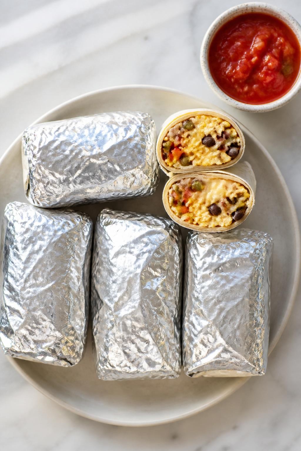 Photo prompt: Realistic top-down editorial food photography of six flour tortilla breakfast burritos wrapped in foil, one cut in half to show scrambled eggs, black beans, bell peppers, onions, and melted pepper jack cheese. A side of salsa in a small bowl. Bright morning light, portable freezer breakfast mood, no people, no hands, no text, no watermarks, no props with writing.