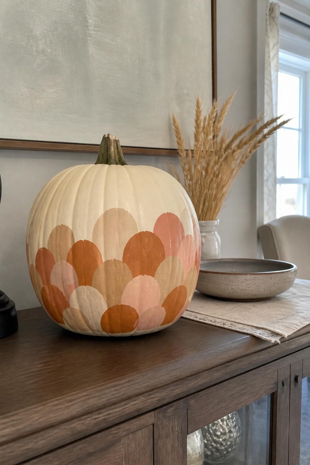 Horizontal 16:9 natural iPhone photograph of a real cream pumpkin painted with an easy overlapping scallop pattern around the lower two-thirds in muted peach, burnt orange, and warm beige, visible curved brushstrokes, displayed on a dining room sideboard beside a ceramic bowl, folded linen runner, and dried wheat stems, natural window light, genuine home atmosphere, realistic matte acrylic paint, no artificial shine, no text.