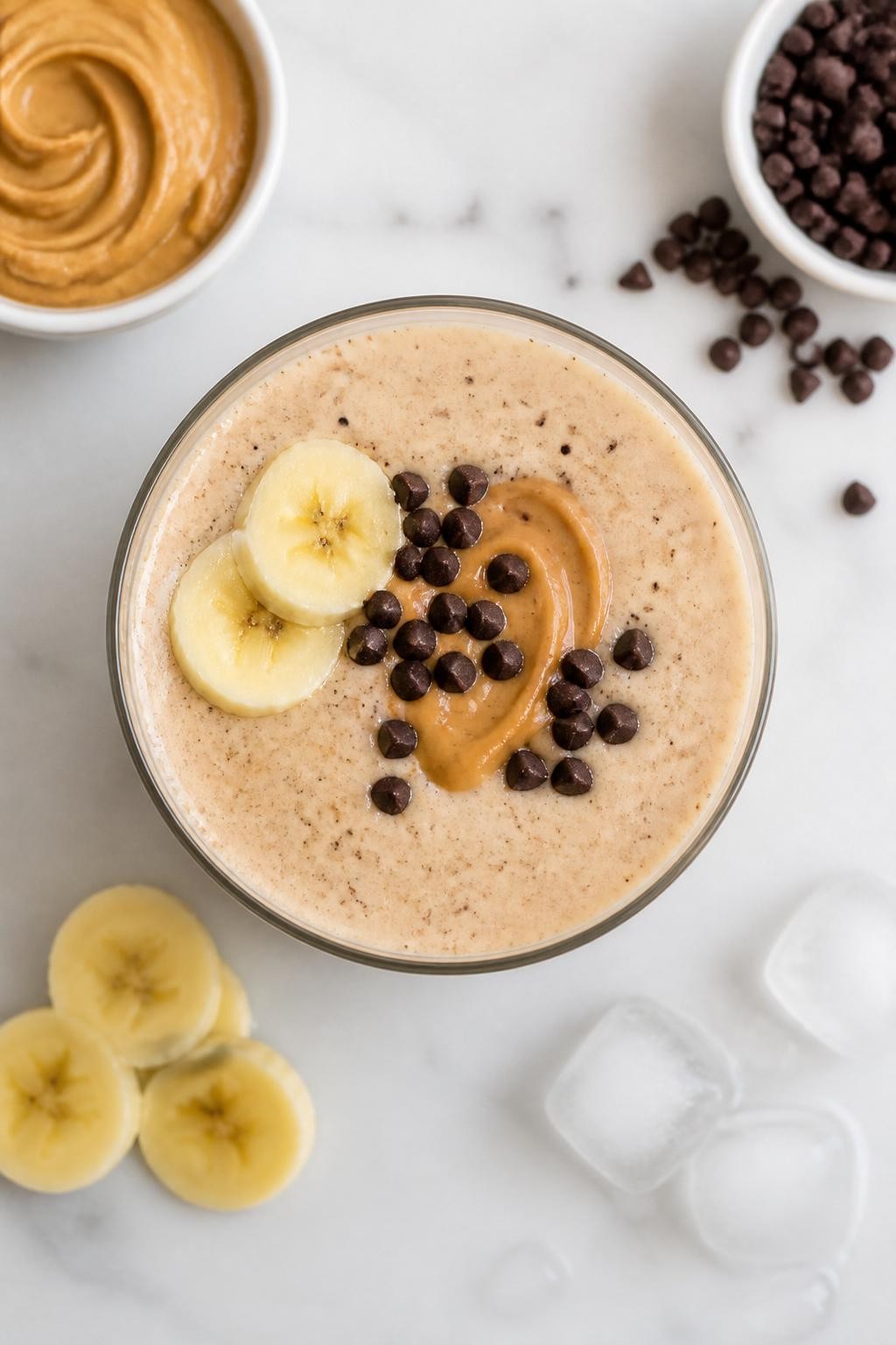 Realistic top-down editorial food photography of a peanut butter chocolate chip smoothie in a clear glass on a clean white marble countertop. The smoothie is creamy beige with tiny dark chocolate flecks, thick and smooth, styled with peanut butter in a plain dish, mini dark chocolate chips, banana slices, and a few ice cubes nearby. Soft natural daylight, clean dessert smoothie styling, high detail, realistic smoothie texture, no people, no hands, no text, no watermarks, no props with writing.
