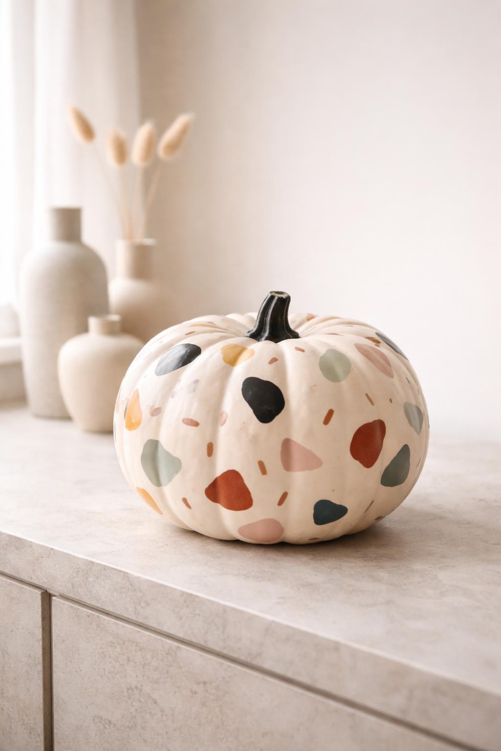 Image Prompt: A pale beige pumpkin decorated in a modern terrazzo pattern, scattered irregular painted chips in terracotta, charcoal, mustard, sage, dusty pink, cream, and muted blue, short matte-black stem, clean contemporary styling on a light stone console with simple ceramic vases, soft natural window light, realistic DSLR photography, crisp painted shapes, no people, no text.
