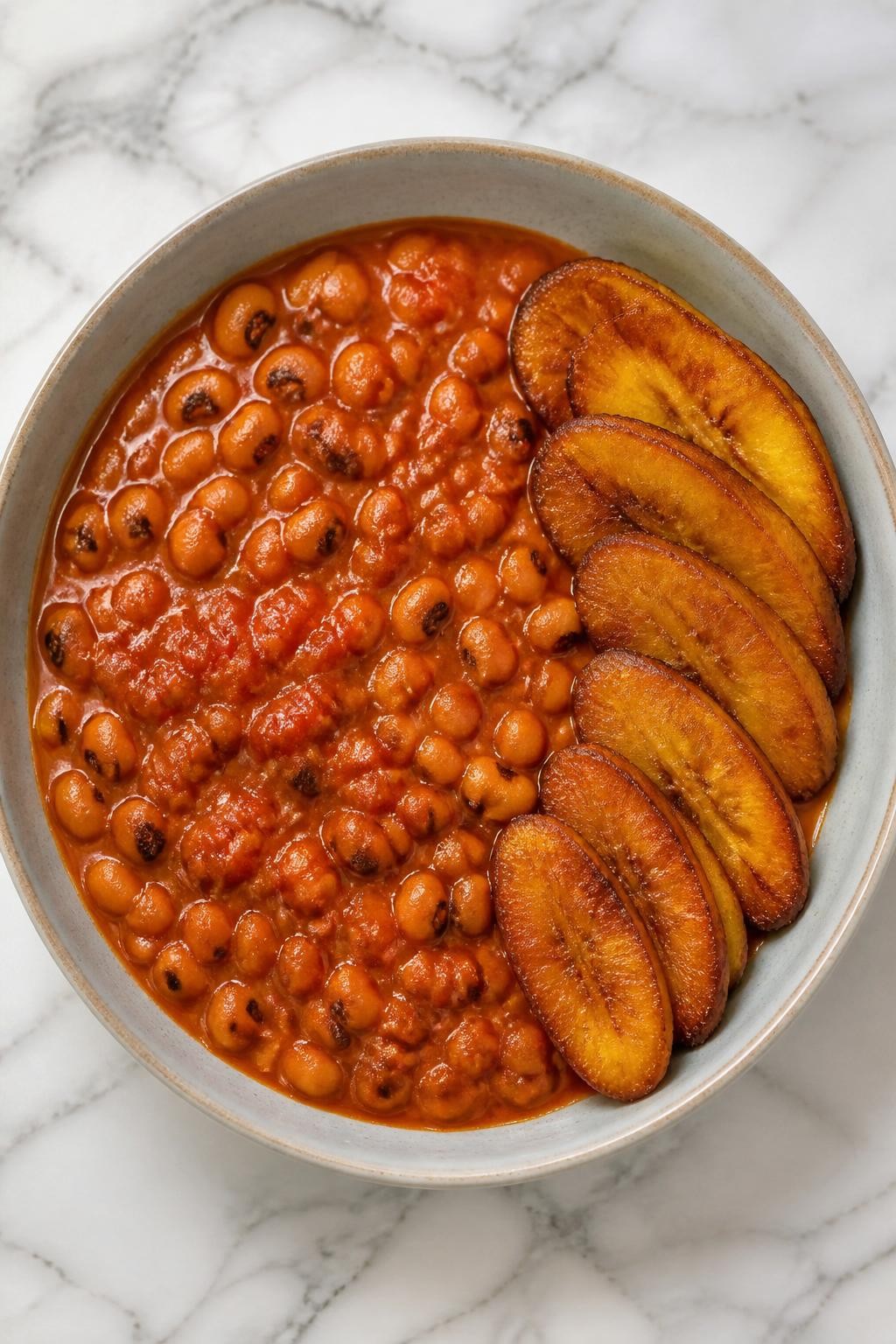 Image Prompt: Top-down realistic food photography of vegan red red on a white marble countertop, black-eyed beans in a rich red tomato stew served with golden fried plantains, warm natural daylight, comforting West African dinner mood, no people, no hands, no text, no watermarks, no props with writing.