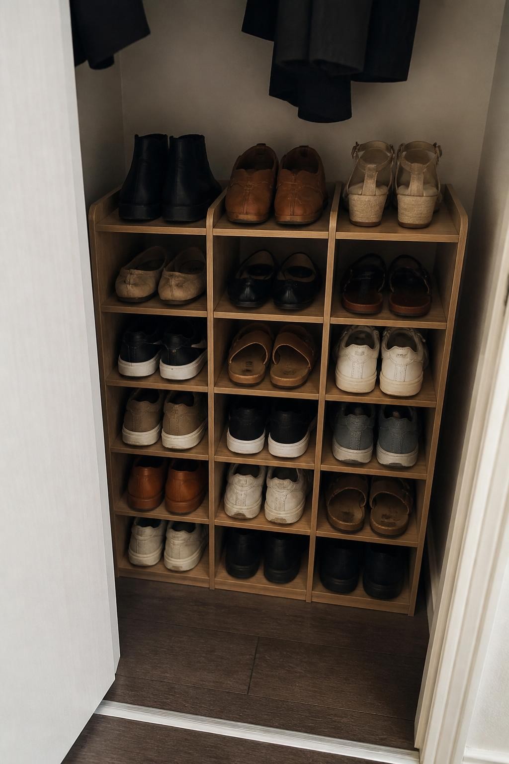 Image prompt: Small closet floor fitted with narrow stackable wooden shoe cubbies, each compartment holding one pair of everyday shoes including loafers, sneakers, flats, sandals, and ankle boots, photographed in modern iPhone photo quality, indirect daylight, believable scuffs and natural shoe textures, practical apartment closet, clean but not staged like a showroom, neutral walls, no people, no brand logos, no watermarks on images.