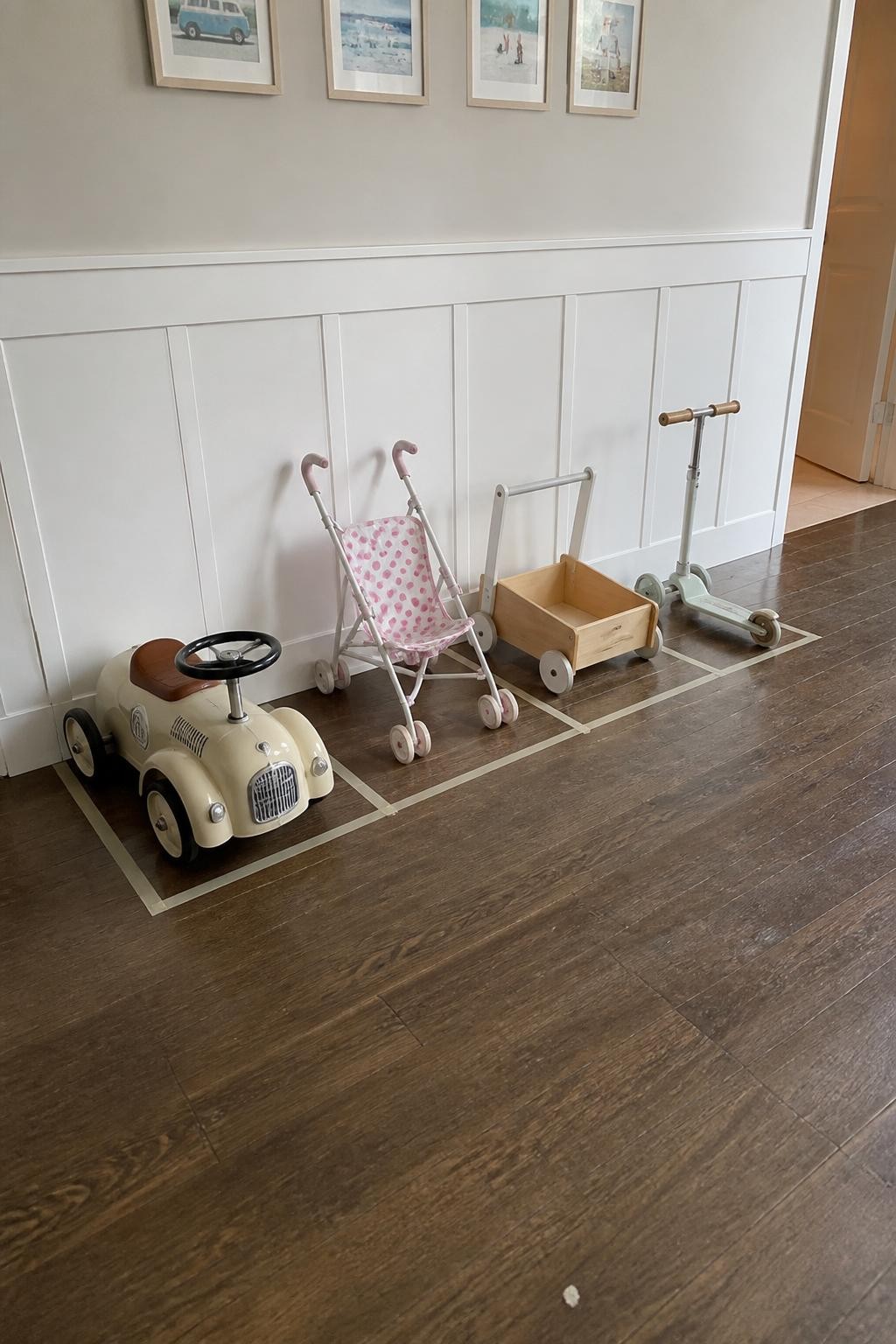Image prompt: Playroom wall with subtle floor tape marking individual parking spaces for a small ride-on car, toy stroller, wooden push cart, and child-sized scooter, photographed with realistic iPhone photo quality, bright natural daylight, believable scuffs on the toys and floor, practical family organization system, clear walking path, no children visible, no logos, no watermarks on images.
