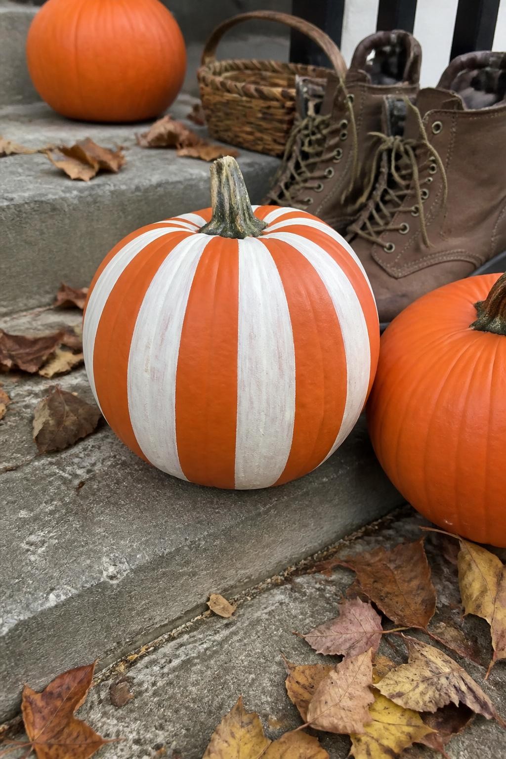Horizontal 16:9 realistic iPhone photograph of a real orange pumpkin painted with wide warm white vertical stripes following the natural ridges from stem to base, visible handmade brush marks, sitting on weathered outdoor steps with plain pumpkins, brown boots, a small basket, and fallen leaves, soft overcast fall daylight, authentic residential setting, natural shadows, no professional lighting, no text.