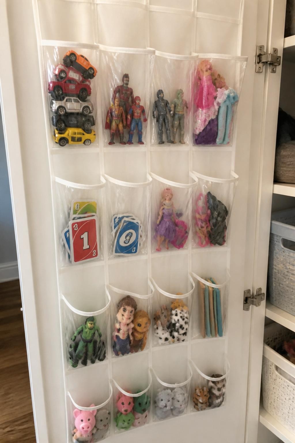 Image prompt: Inside of a playroom cupboard door fitted with a clear pocket organizer holding small toy cars, action figures, card games, doll accessories, and finger puppets, photographed with natural iPhone photo quality, soft side light, realistic mixed toy sizes and slightly imperfect arrangement, ordinary family home storage, no children, no visible logos, no watermarks on images.