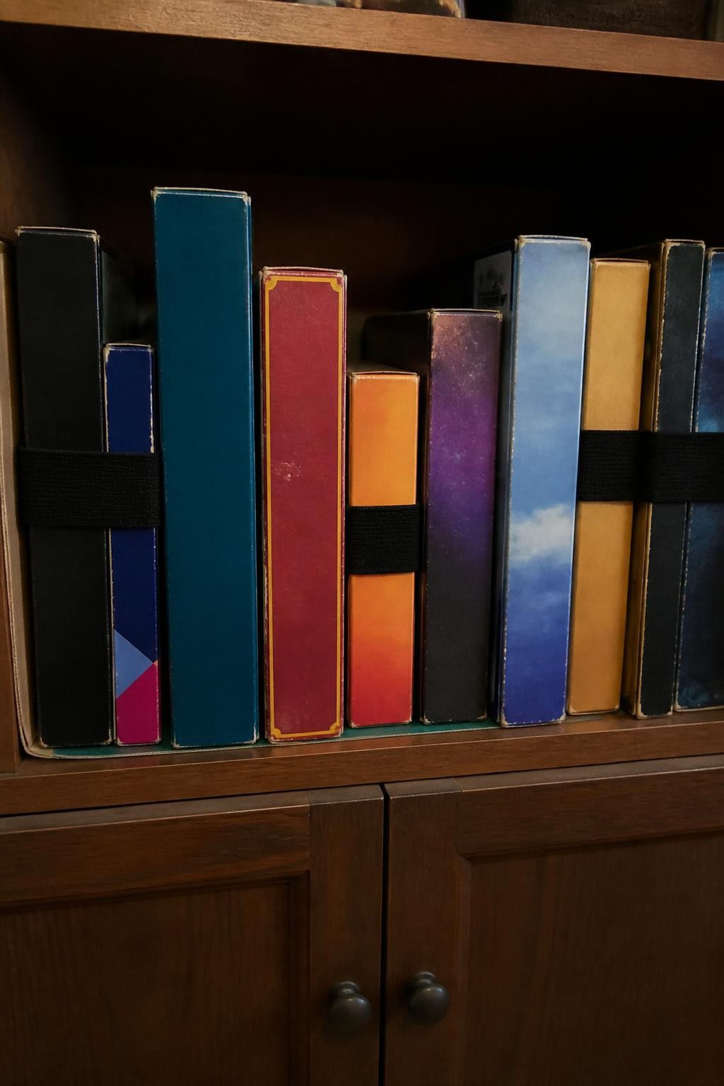 Image prompt: Family cupboard shelf with board games stored vertically like books between sturdy dividers, several boxes secured with wide elastic bands, photographed with natural iPhone photo quality, warm evening light, believable worn box corners and mixed game sizes, ordinary home storage, no readable commercial titles or logos, no people, no watermarks on images.