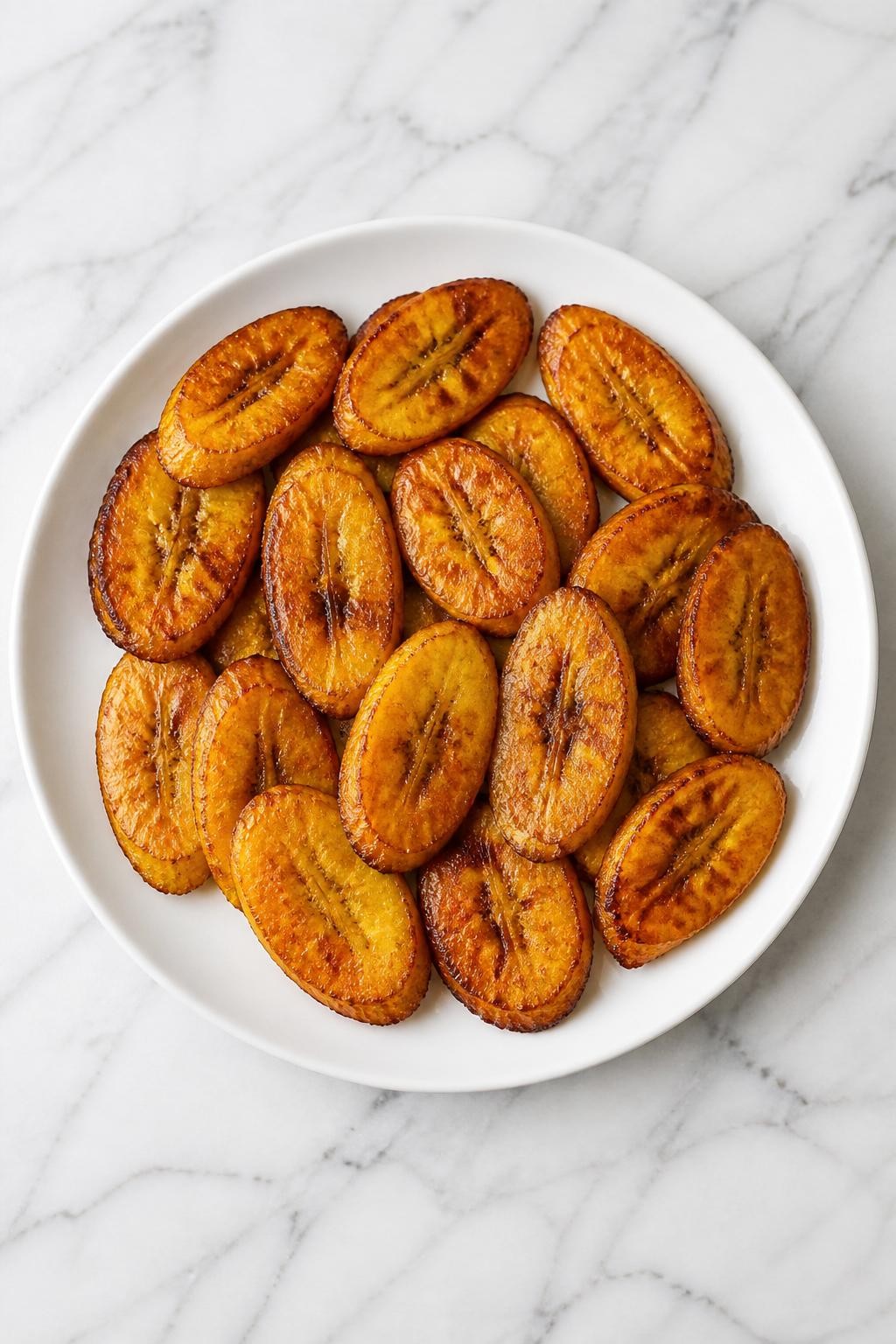 Image Prompt: Top-down realistic editorial food photography of golden fried plantain slices on a clean white marble countertop, caramelized ripe plantain pieces arranged neatly on a simple white plate, bright natural daylight, warm cheerful homemade African side dish mood, clean composition, no people, no hands, no text, no watermarks, no props with writing.